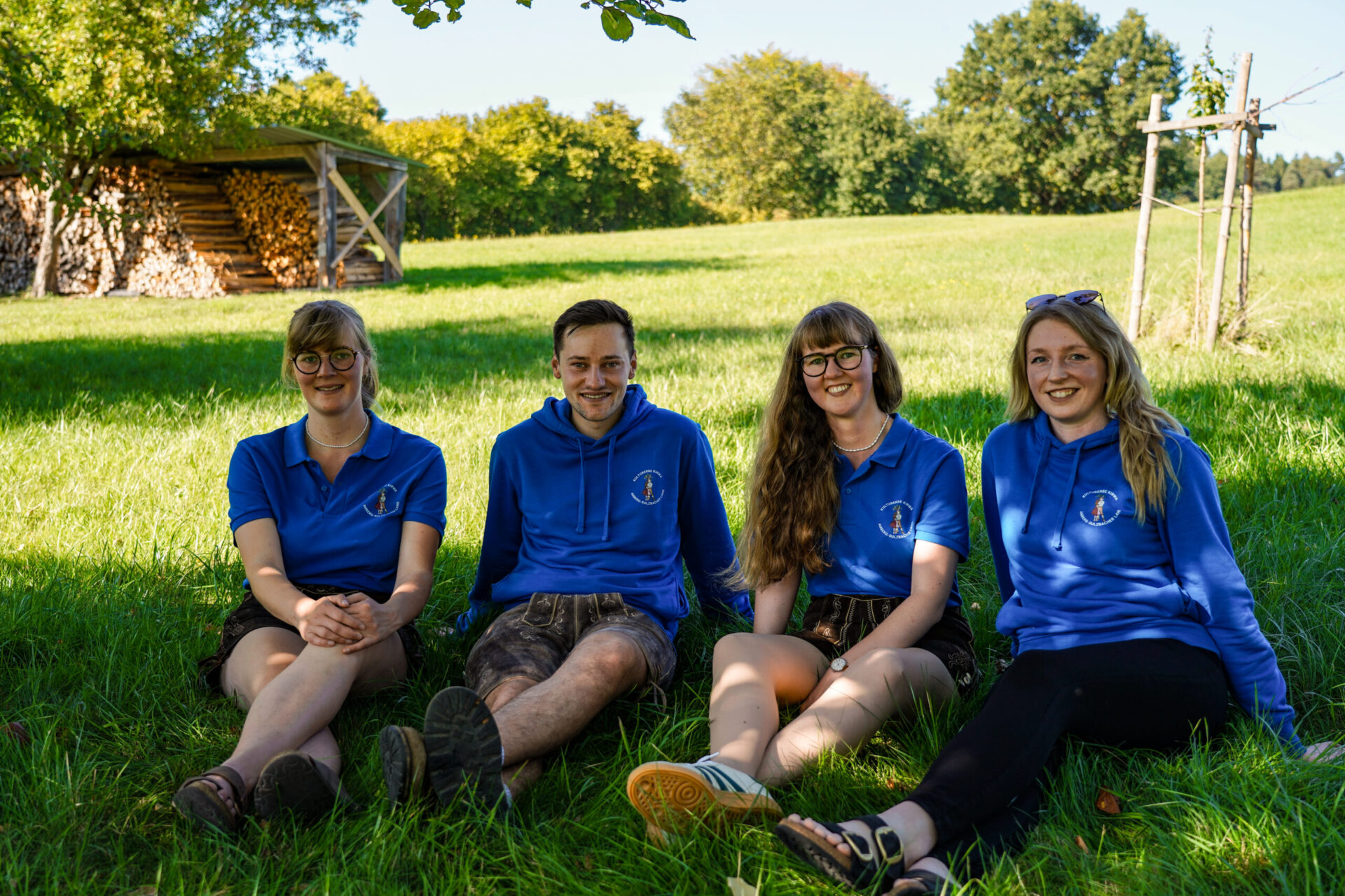 3 Frauen und ein Mann sitzen auf einer Wiese und tragen blaue Shirts und Hoodies.