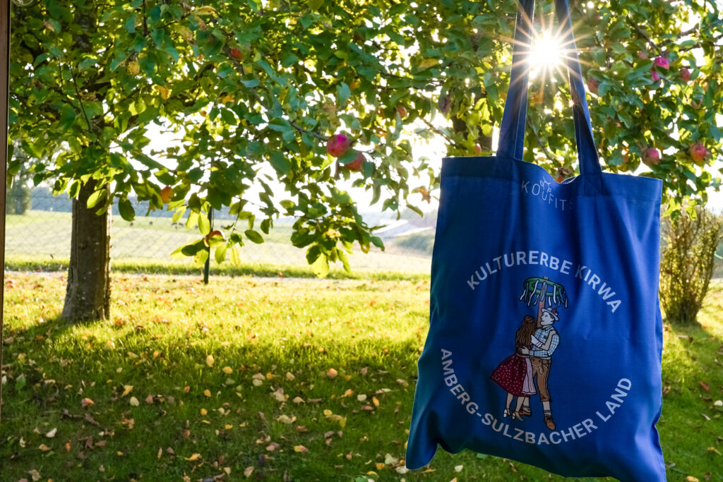 Eine blaue Baumwolltasche hängt an einem Apfelbaum, durch die Henkel scheint die Sonne.